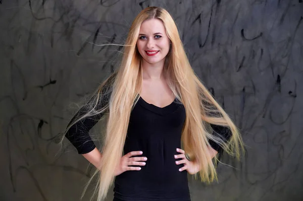 Conceito de uma jovem mulher loira com um penteado chique. Retrato de uma menina bonita em uma camiseta preta com cabelo longo e bonito e ótima maquiagem. Sorrindo, mostrando emoções em um fundo cinza . — Fotografia de Stock