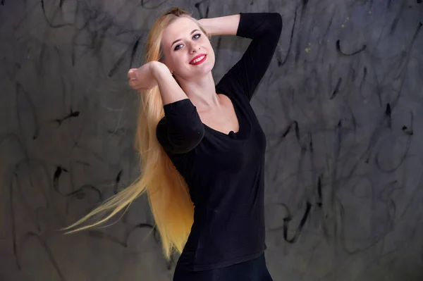 Conceito de uma jovem mulher loira com um penteado chique. Retrato de uma menina bonita em uma camiseta preta com cabelo longo e bonito e ótima maquiagem. Sorrindo, mostrando emoções em um fundo cinza . — Fotografia de Stock