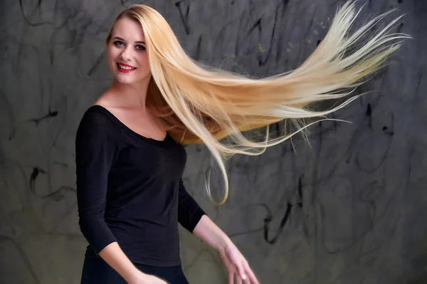 Conceito de uma jovem mulher loira com um penteado chique. Retrato de uma menina bonita em uma camiseta preta com cabelo longo e bonito e ótima maquiagem. Sorrindo, mostrando emoções em um fundo cinza . — Fotografia de Stock