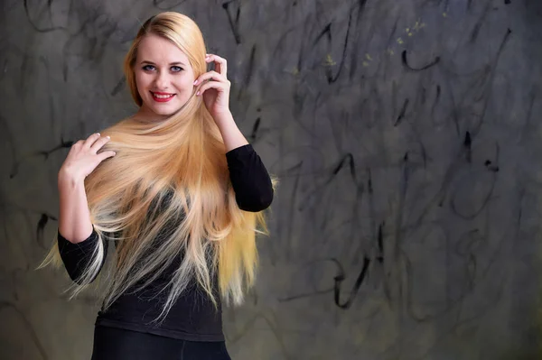 Concepto de una joven rubia con un peinado chic. Retrato de una linda chica en una camiseta negra con pelo largo y hermoso y gran maquillaje. Sonriendo, mostrando emociones sobre un fondo gris . — Foto de Stock