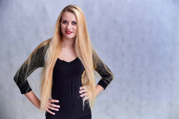 El concepto de una chica rubia con un peinado chic. Retrato de una linda chica en una camiseta negra con pelo largo y hermoso y gran maquillaje. Sonriendo, mostrando emociones sobre un fondo gris claro . — Foto de Stock