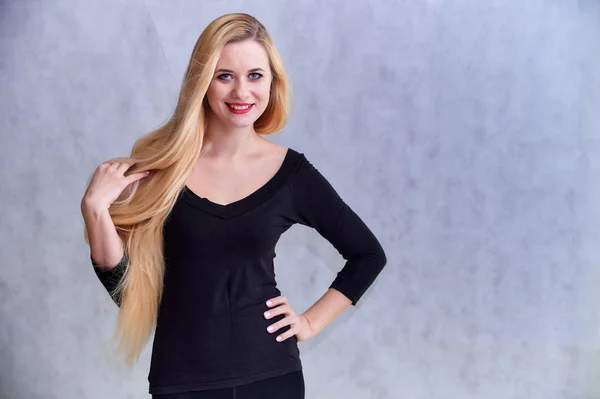 El concepto de una chica rubia con un peinado chic. Retrato de una linda chica en una camiseta negra con pelo largo y hermoso y gran maquillaje. Sonriendo, mostrando emociones sobre un fondo gris claro . — Foto de Stock