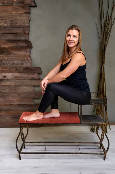 Portrait of a pretty fitness blonde girl in a tracksuit on an alternative background in vintage interior. Cute looks and smiles at the camera, sitting on a stand straight. Loft style concept. — Stock Photo, Image
