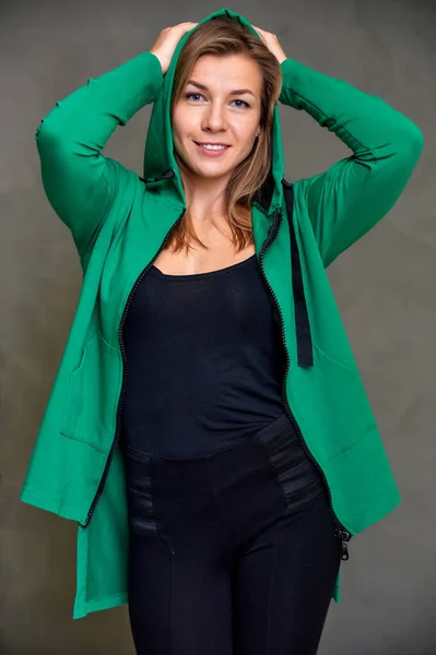 Portret van een mooi fitness blond meisje in een zwart en groen sportpak op een grijze achtergrond in vintage interieur. Schattige blikken met een glimlach naar de camera, rechtop staand. Loft stijl concept. — Stockfoto