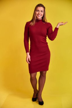 Sarı arka planda bordo bir elbise içinde güzel bir gülümsemesi olan hoş, formda, sarışın bir kızın tam boy dikey portresi. Kameraya bakıyor, dik duruyor..