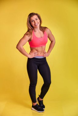 Güzel bir fitness sarışın kızın dikey portresi güzel bir gülümseme, mükemmel dişler ve sarı arka planda eşofman giymiş bir vücut geliştirme figürü. Kameraya bakıyor, dik duruyor..