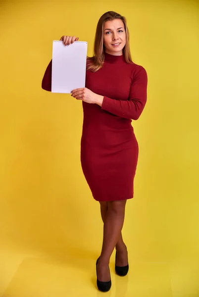 Retrato vertical de cuerpo entero de una bonita chica rubia con una hermosa sonrisa en un vestido borgoña con una carpeta en las manos sobre un fondo amarillo. Lindas miradas a la cámara, de pie recta . — Foto de Stock