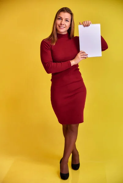 Retrato vertical de cuerpo entero de una bonita chica rubia con una hermosa sonrisa en un vestido borgoña con una carpeta en las manos sobre un fondo amarillo. Lindas miradas a la cámara, de pie recta . — Foto de Stock