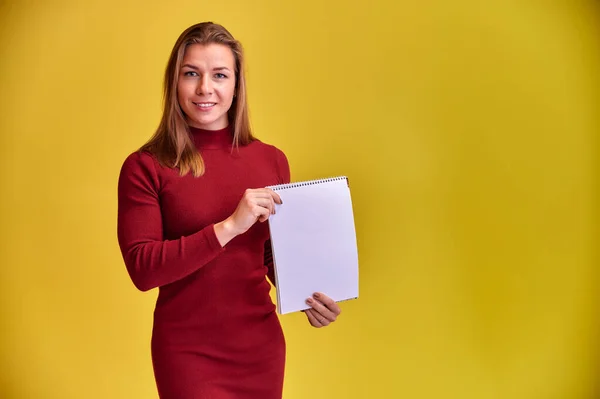 Retrato de una bonita chica rubia con una hermosa sonrisa y excelentes dientes en un vestido borgoña con una carpeta en las manos sobre un fondo amarillo. Lindas miradas a la cámara, de pie recta . — Foto de Stock