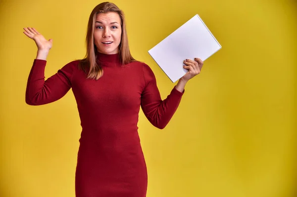 Ritratto di una bella ragazza bionda con un bel sorriso e denti eccellenti in un vestito bordeaux con una cartella tra le mani su uno sfondo giallo. Carino guarda la fotocamera, in piedi dritto . — Foto Stock