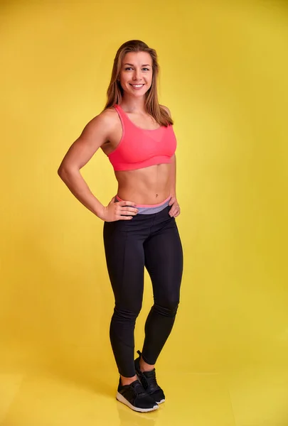 Das vertikale Porträt einer hübschen Fitness-Blondine mit einem schönen Lächeln, hervorragenden Zähnen und einer Bodybuilderfigur im Trainingsanzug auf gelbem Hintergrund. Niedliche Blicke in die Kamera, gerade stehend. — Stockfoto