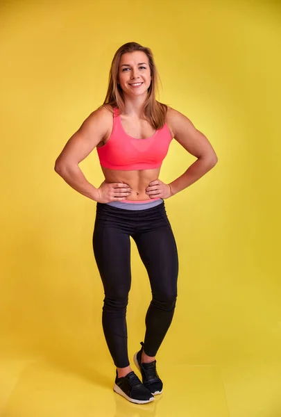 Das vertikale Porträt einer hübschen Fitness-Blondine mit einem schönen Lächeln, hervorragenden Zähnen und einer Bodybuilderfigur im Trainingsanzug auf gelbem Hintergrund. Niedliche Blicke in die Kamera, gerade stehend. — Stockfoto