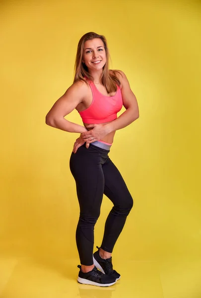Retrato vertical de uma linda garota loira fitness com um belo sorriso, dentes excelentes e uma figura de fisiculturista em um traje de treino em um fundo amarelo. Bonito olha para a câmera, em pé em linha reta . — Fotografia de Stock
