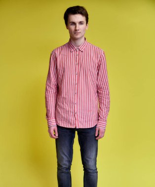 Pembe gömlekli, sarı arka planda kot pantolonlu, kısa saçlı yakışıklı bir gencin dikey portresi. Kameraya bakıyor, dik duruyor..