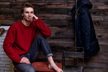 Yakışıklı bir gencin portresi. Eskimiş ahşap arka planda bordo süveter giyen kısa saçlı beyaz. Kürsüde otururken kameraya tatlı tatlı bakıyor. Çatı katı biçimi.