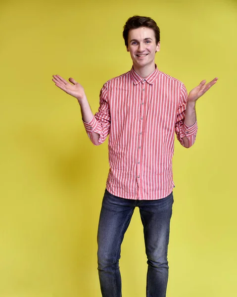 Vertikální portrét pohledného mladíka s krátkým účesem a širokým úsměvem v růžové košili a džínách na žlutém pozadí. Roztomilý pohled na kameru, stojí rovně. — Stock fotografie