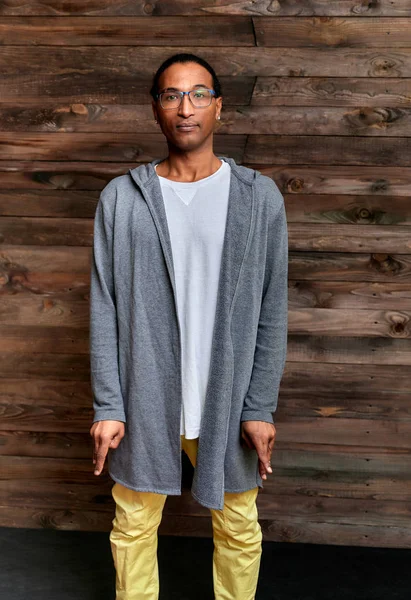 Pionowy Portret Młody mężczyzna w krótkowłosych afro-amerykańskich okularach w zwykłych ubraniach na drewnianym tle w stylu loftowym. Stojąc tuż przed kamerą — Zdjęcie stockowe