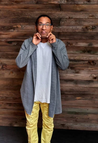Retrato vertical Hombre joven con gafas afroamericanas de pelo corto en ropa ordinaria sobre un fondo de madera vintage en el interior de una casa en un estilo loft. De pie justo delante de la cámara —  Fotos de Stock