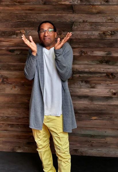 Retrato vertical Hombre joven con gafas afroamericanas de pelo corto en ropa ordinaria sobre un fondo de madera vintage en el interior de una casa en un estilo loft. De pie justo delante de la cámara —  Fotos de Stock