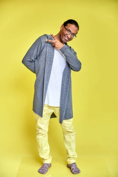 Retrato vertical de un joven alegre con gafas afroamericanas con un corte de pelo corto en ropa ordinaria sobre un fondo amarillo en toda su longitud. De pie justo delante de la cámara —  Fotos de Stock