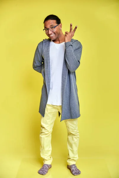 Vertikalt porträtt av en glad ung man i glasögon afro-amerikan med en kort frisyr i vanliga kläder på en gul bakgrund i full längd. Stående framför kameran — Stockfoto