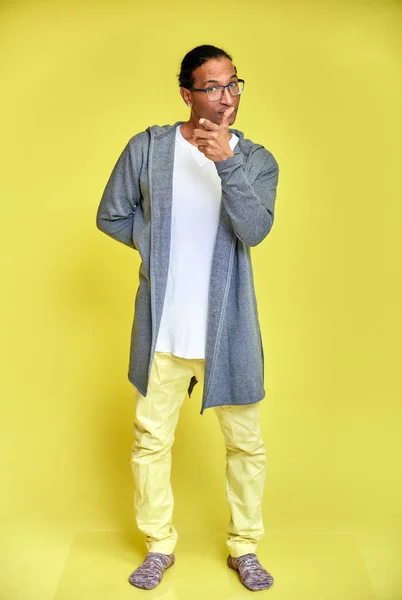 Retrato vertical de un joven alegre con gafas afroamericanas con un corte de pelo corto en ropa ordinaria sobre un fondo amarillo en toda su longitud. De pie justo delante de la cámara —  Fotos de Stock