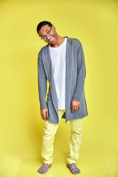 Retrato vertical de un joven alegre con gafas afroamericanas con un corte de pelo corto en ropa ordinaria sobre un fondo amarillo en toda su longitud. De pie justo delante de la cámara —  Fotos de Stock