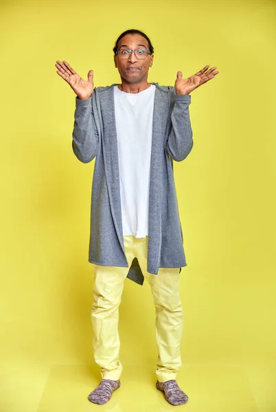 Retrato vertical de un joven alegre con gafas afroamericanas con un corte de pelo corto en ropa ordinaria sobre un fondo amarillo en toda su longitud. De pie justo delante de la cámara —  Fotos de Stock