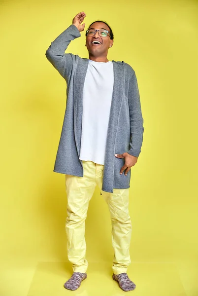 Retrato vertical de un joven alegre con gafas afroamericanas con un corte de pelo corto en ropa ordinaria sobre un fondo amarillo en toda su longitud. De pie justo delante de la cámara —  Fotos de Stock