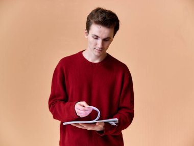 Kısa saçlı, beyaz dişli, pembe arka planda bordo süveterli genç bir beyaz adamın portresi. Elinde bir klasörle kameranın önünde el ele tutuşup konuşmak..