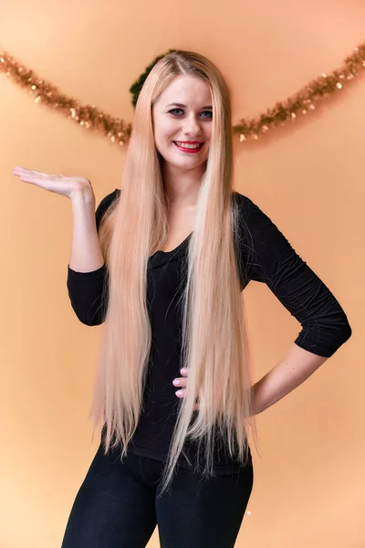 Retrato de una linda chica en una camiseta negra con pelo largo y hermoso y gran maquillaje. Concepto de una joven rubia con decoración de Año Nuevo. Sonriendo, mostrando emociones sobre un fondo rosado . — Foto de Stock