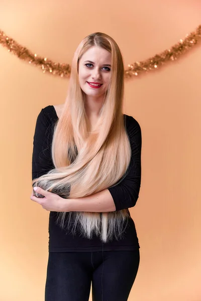 Portrait of a cute girl in a black T-shirt with long beautiful hair and great makeup. Concept of a young blonde woman with New Year's decor. Smiling, showing emotions on a pink background. — ストック写真