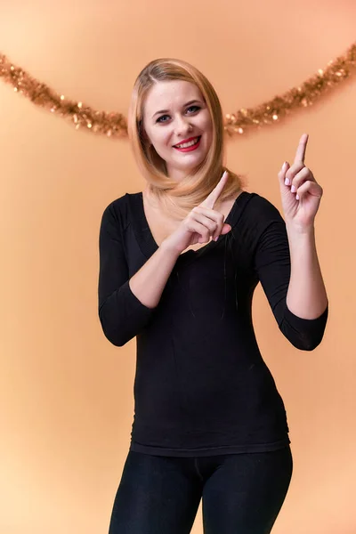Retrato de uma menina bonita em uma camiseta preta com cabelo longo e bonito e ótima maquiagem. Conceito de uma jovem loira com decoração de Ano Novo. Sorrindo, mostrando emoções em um fundo rosa . — Fotografia de Stock