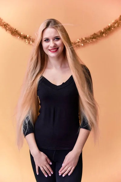 Retrato de una linda chica en una camiseta negra con pelo largo y hermoso y gran maquillaje. Concepto de una joven rubia con decoración de Año Nuevo. Sonriendo, mostrando emociones sobre un fondo rosado . — Foto de Stock