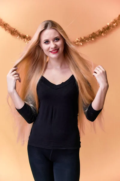 Retrato de una linda chica en una camiseta negra con pelo largo y hermoso y gran maquillaje. Concepto de una joven rubia con decoración de Año Nuevo. Sonriendo, mostrando emociones sobre un fondo rosado . — Foto de Stock
