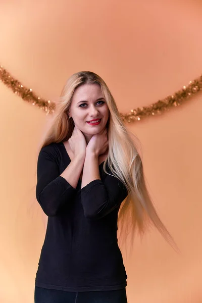 Portrait of a cute girl in a black T-shirt with long beautiful hair and great makeup. Concept of a young blonde woman with New Year's decor. Smiling, showing emotions on a pink background. — ストック写真