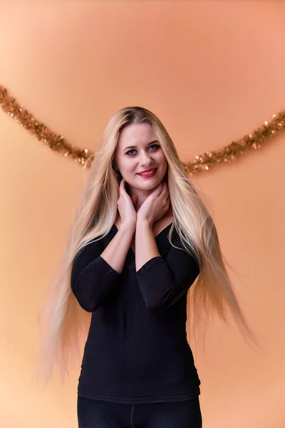 Portrait of a cute girl in a black T-shirt with long beautiful hair and great makeup. Concept of a young blonde woman with New Year's decor. Smiling, showing emotions on a pink background. — ストック写真