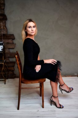 Alternatif bir iç mekanda, sandalyede oturan, siyah elbiseli, ince, sarışın bir kadının tam boy sanat portresi. Retro, cazibe, romantizm kavramı. Yan görünüm