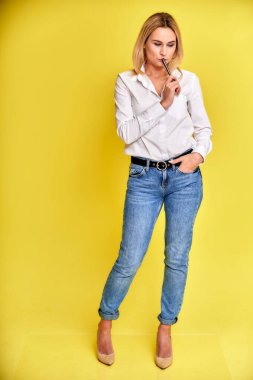 Sarı arka planda duran mavi kotlu ve beyaz gömlekli, gülümseyen sarışın bir kadının tam boy sanat portresi. Hem de kameranın önünde..