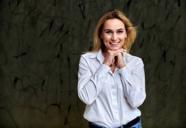 Beyaz tişörtlü, gri arka planda uçan saçlı, gülümseyen sarışın bir kadının portresi. Hem de kameranın önünde..