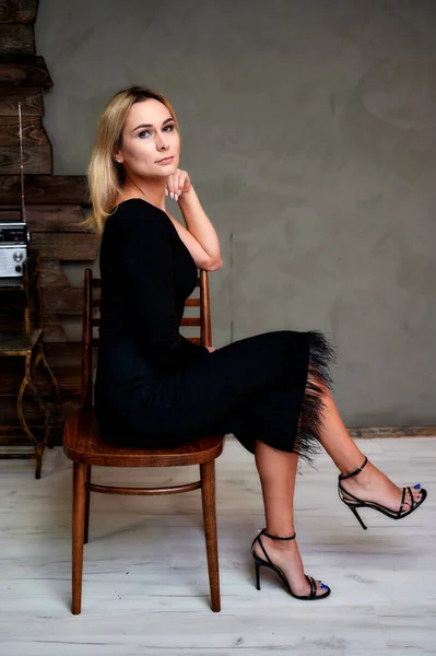 Retrato de arte completo de uma mulher loira muito esbelta em um vestido preto sentado em uma cadeira em um interior alternativo. O conceito de retro, glamour, romance. Vista lateral — Fotografia de Stock
