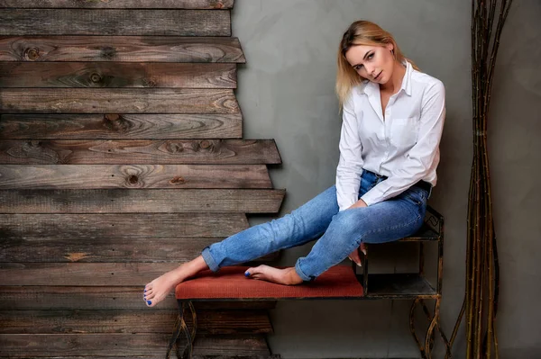 Full-length art portret van een mooie blonde vrouw in blauwe jeans en een wit shirt zittend op een standaard in een alternatief interieur. Het concept van retro, glamour, romantiek. — Stockfoto