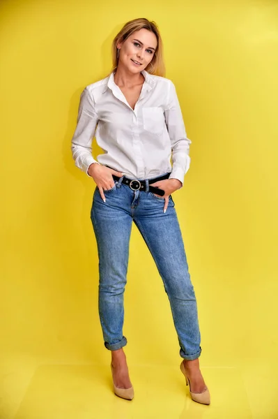 Полнометражный художественный портрет красивой улыбающейся блондинки в синих джинсах и белой рубашке стоит на жёлтом фоне. Прямо перед камерой с эмоциями . — стоковое фото