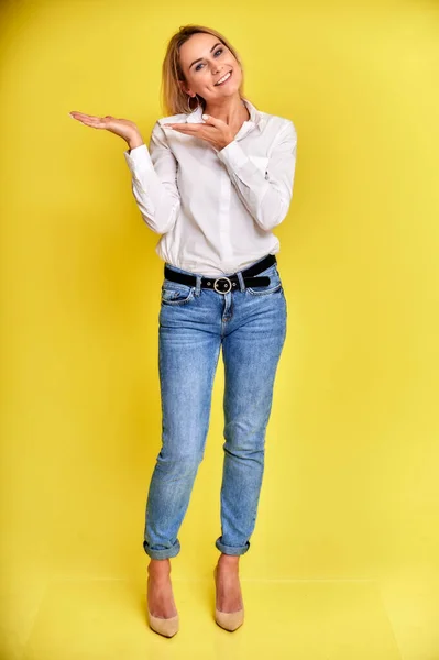 Retrato de arte completo de uma mulher loira muito sorridente em jeans azuis e uma camisa branca fica em um fundo amarelo. Bem na frente da câmera com emoções . — Fotografia de Stock