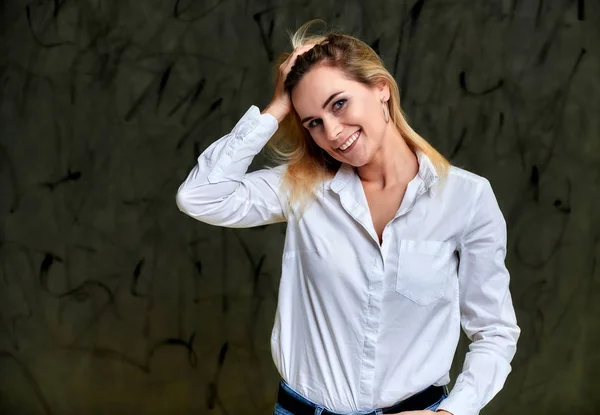 Retrato de una hermosa mujer rubia sonriente con una camisa blanca con el pelo volador de pie sobre un fondo alternativo gris. Justo en frente de la cámara con emociones . — Foto de Stock