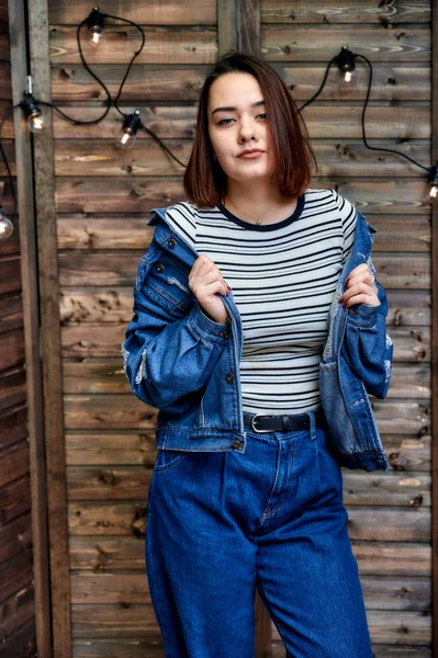 Portrait of a pretty brunette girl in denim suit on wooden background in alternative interior. Standing right in front of the camera in various poses. — Stock Photo, Image