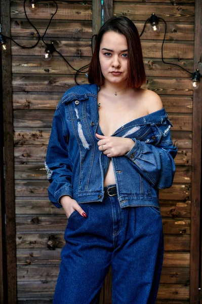 Portrait of a pretty brunette girl in denim suit on wooden background in alternative interior. Standing right in front of the camera in various poses. — 스톡 사진