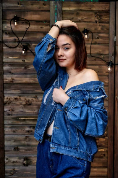 Portrait of a pretty brunette girl in denim suit on wooden background in alternative interior. Standing right in front of the camera in various poses. — 스톡 사진