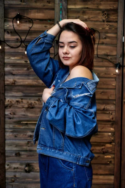 Portrait of a pretty brunette girl in denim suit on wooden background in alternative interior. Standing right in front of the camera in various poses. — Stock Photo, Image