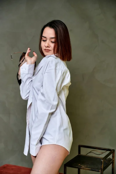 Portrait of a pretty brunette Caucasian girl in a white shirt on a gray background in an alternative interior. Standing right in front of the camera in various poses. — 스톡 사진